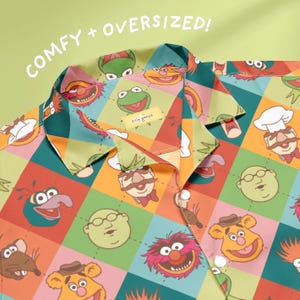 May include: A colorful, oversized button-down shirt featuring a patchwork design with various Muppet characters. The shirt has a collar and buttons, with the words "COMFY + OVERSIZED!" in white text. The shirt is made of a soft, comfortable material.