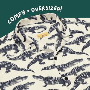 May include: Cream-colored button-down shirt with a repeating pattern of navy blue alligators. The shirt has a classic collar and black buttons. Text on the shirt reads "COMFY + OVERSIZED!" and a small tag reads "SILLY GOOSE".