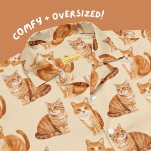 May include: A beige button-down shirt featuring an all-over print of orange tabby cats. The collar is visible, and a small yellow tag inside the neck reads "SILLY GOOSE WORKSHOP & WARES". The text "COMFY + OVERSIZED!" appears in white above the shirt.