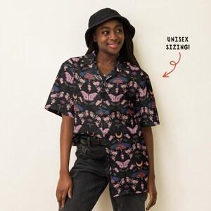 May include: A black short-sleeved button-down shirt featuring a repeating pattern of pink and blue moths, small flowers, and crescent moons. The shirt includes a collar and a front button closure. Text on the right side reads UNISEX SIZING!
