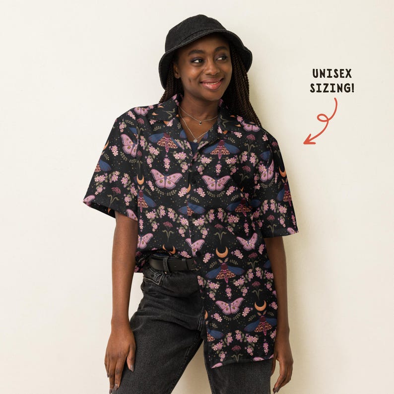 May include: A black short-sleeved button-down shirt featuring a repeating pattern of pink and blue moths, small flowers, and crescent moons. The shirt includes a collar and a front button closure. Text on the right side reads UNISEX SIZING!