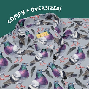May include: A button-down shirt with a gray background and a pattern of watercolor pigeons. The shirt has a collar and black buttons. The text "COMFY + OVERSIZED!" is in white letters at the top. A small tag reads "SILLY GOOSE".