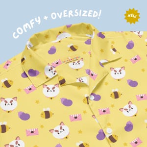 May include: A yellow button-down shirt featuring a repeating pattern of white cat faces, purple eggplants, bees, pink envelopes, and small yellow stars. The collar is open, and the inner label reads Silly Goose Workshop & Wares. Text reads COMFY + OVERSIZED!