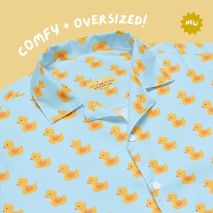 May include: Light blue button-up shirt with a repeating pattern of yellow rubber duckies. The shirt has a collar and buttons down the front. The text "COMFY + OVERSIZED!" is at the top, and the text "NEW" is in the upper right corner.