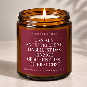 Könnte beinhalten: Eine brennende Kerze in einem bernsteinfarbenen Glas mit einem dunkelroten Etikett. Das Etikett trägt den Text: INDIGOFOXCANDLES, UNS ALS ANGESTELLTE ZU HABEN, IST DAS EINZIGE GESCHENK, DAS DU BRAUCHST, und HANDMADE SCENTED SOY WAX CANDLE in Weiß.