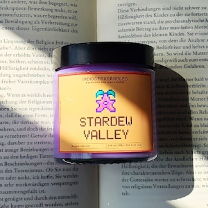 Op de afbeelding: Een paarse kaars met een zwart deksel, met het opschrift "STARDEW VALLEY" en een gepixeld karakter. De kaars staat op een open boek met tekst.