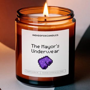 Könnte beinhalten: Eine braune Glaskerze mit einem weißen Etikett, auf dem "INDIGOFOXCANDLES The Mayor's Underwear" steht. Das Etikett zeigt ein pixeliertes Bild von lila Unterwäsche.