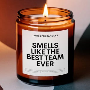 Può includere: Una candela accesa in un barattolo cilindrico di vetro ambrato con un'etichetta bianca. L'etichetta riporta il testo INDIGOFOXCANDLES in alto e le parole SMELLS LIKE THE BEST TEAM EVER in grassetto nero, con del testo più piccolo in basso.