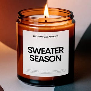 Könnte beinhalten: Eine brennende Kerze in einem durchsichtigen Glas mit einem braunen Etikett mit der Aufschrift "SWEATER SEASON". Die Kerze ist von Indigo Fox Candles. Der Hintergrund ist eine Mischung aus Braun, Weiß und Grau.