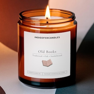 May include: A lit candle in an amber glass jar with a white label reading INDIGOFOXCANDLES, Old Books, Teakwood, Oak, Sandalwood, and featuring a small book icon. The candle has a single wick and is filled with wax, emitting a warm, bright flame.