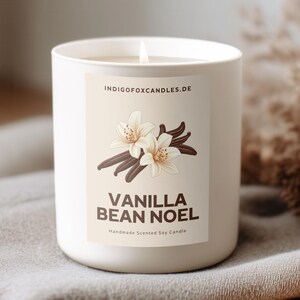 Op de afbeelding: Een witte kaars met een brandende lont in een keramische houder met het label "Vanilla Bean Noel". Het label toont vanillebloemen en -bonen. De kaars staat op een grijze deken met een franje, met gedroogde planten op de achtergrond.