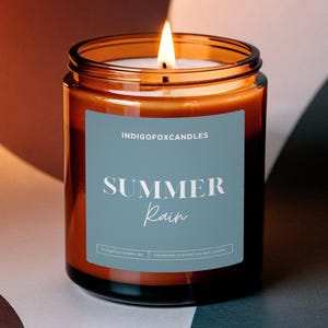 Op de afbeelding: Een brandende kaars in een heldere glazen pot met een bruine tint. De kaars heeft een blauw etiket met de tekst "SUMMER Rain" en "INDIGOFoxCandles". De achtergrond is een mix van bruine, witte en zwarte vormen.
