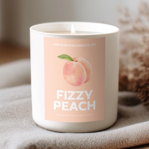 Könnte beinhalten: Eine brennende weiße Kerze mit einem Etikett, das eine Pfirsich-Illustration und die Worte "Fizzy Peach" zeigt. Die Kerze steht auf einer grauen Decke, mit getrockneten Pflanzen und einem Weidenkorb im Hintergrund. Es ist ein Dekorationsartikel.