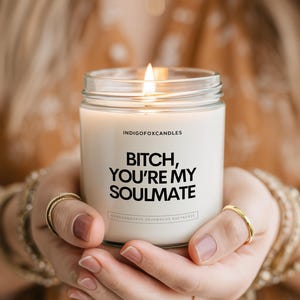 Könnte beinhalten: Eine Kerze im klaren Glasgefäß mit einem weißen Etikett, das den schwarzen Text "INDIGOFORCANDLES" und "BITCH, YOU'RE MY SOULMATE" in fetten Großbuchstaben zeigt. Die Kerze enthält weißes Wachs und hat einen einzelnen brennenden Docht.