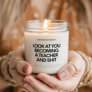 Könnte beinhalten: Eine Kerze im klaren Glasgefäß mit weißem Wachs und brennendem Docht. Das Etikett trägt den schwarzen Text INDIGOFOKCANDLES, LOOK AT YOU BECOMING A TEACHER AND SHIT sowie kleineren Text darunter. Die Kerze befindet sich in einem zylindrischen Glasbehälter.