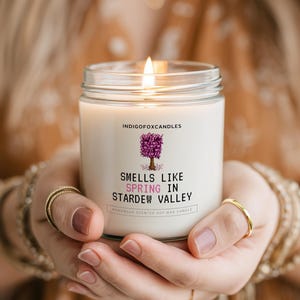 Op de afbeelding: Een brandende kaars in een heldere glazen pot. De kaars heeft een wit etiket met de tekst "Smells Like Spring in Stardew Valley" en een paarse boomafbeelding. De kaars wordt met beide handen vastgehouden.