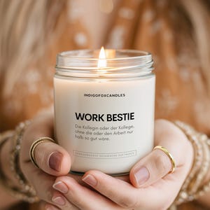 Op de afbeelding: Een brandende kaars in een heldere glazen pot. De kaars heeft een wit etiket met de woorden "WORK BESTIE" en extra tekst. De kaars wordt voor een wazige achtergrond gehouden.