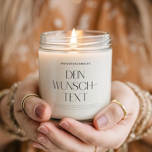 Könnte beinhalten: Eine brennende weiße Kerze in einem durchsichtigen Glas. Auf der Kerze steht der Text "DEIN WUNSCH-TEXT". Die Kerze wird mit beiden Händen gehalten, wobei goldene Ringe und Armbänder zu sehen sind. Der Hintergrund ist unscharf.