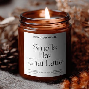 Puede incluir: Una vela encendida en un tarro de vidrio marr&oacute;n con una etiqueta blanca que dice "Smells like Chai Latte". La vela est&aacute; rodeada de plantas secas y una pi&ntilde;a sobre una superficie gris texturizada. La vela es una vela de cera de soja perfumada hecha a mano.