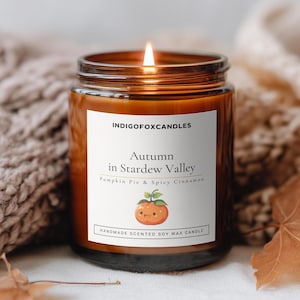 Può includere: Una candela accesa in un barattolo di vetro ambrato con un'etichetta bianca che riporta la scritta INDIGOFOXCANDLES, Autumn in Stardew Valley, Pumpkin Pie & Spicy Cinnamon, e Handmade Scented Soy Wax Candle, con un'illustrazione di zucca.