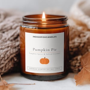 Könnte beinhalten: Eine brennende Sojawachskerze in einem bernsteinfarbenen Glasgefäß mit einem weißen Etikett, das INDIGOFOXCANDLES, Pumpkin Pie, Pumpkin Spice & Salted Caramel und Handmade Scented Soy Wax Candle sowie eine kleine orangefarbene Kürbisgrafik zeigt.