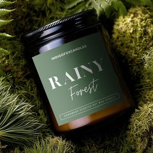 Könnte beinhalten: Ein dunkel bernsteinfarbenes Glas-Kerzenglas mit schwarzem Deckel und grünem Etikett. Das Etikett zeigt den Text INDIGOFOXCANDLES, RAINY Forest und HANDMADE SCENTED SOY WAX CANDLE in weißer und silberner Schrift, umgeben von grünen Farnen.
