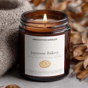 Könnte beinhalten: Eine brennende Kerze in einem braunen Glasgefäß mit einem weißen Etikett, auf dem INDIGOFOXCANDLES, Japanese Bakery, Melon Bread & Creamy Vanilla und Handmade Scented Soy Wax Candle steht, inklusive einer Abbildung eines Melon-Brot-Brötchens.