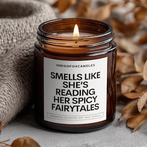 Könnte beinhalten: Eine braune Glasbehälterkerze mit einem weißen Etikett, das den Text INDIGOFOXCANDLES, SMELLS LIKE SHE'S READING HER SPICY FAIRYTALES und HANDMADE SCENTED SOY WAX CANDLE trägt. Die Kerze hat einen brennenden Docht und ist mit Sojawachs gefüllt.