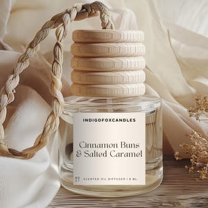 Könnte beinhalten: Ein klarer Glas-Duftöldiffusor mit einem Holzdeckel und einem geflochtenen Seil. Auf dem Etikett steht "Cinnamon Buns & Salted Caramel". Der Diffusor wird auf einer hellen Oberfläche mit einem weichen, neutralen Hintergrund präsentiert.