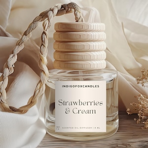 Può includere: Un diffusore di olio profumato in vetro trasparente con coperchio in legno e corda intrecciata. L'etichetta recita "Strawberries & Cream" e "IndigoFoxCandles". Il diffusore è esposto su una superficie di legno con un tessuto morbido dai toni neutri sullo sfondo.