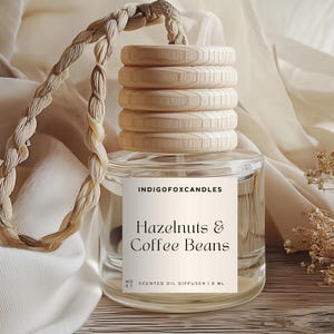 Könnte beinhalten: Ein klarer Glas-Duftöldiffusor mit einem Holzdeckel und einem geflochtenen Seilgriff. Auf dem Etikett steht "Hazelnuts & Coffee Beans" und "IndigoFoxCandles". Der Diffusor hat 1,8 ml und wird auf einer Holzoberfläche mit cremefarbenem Stoffhintergrund präsentiert.