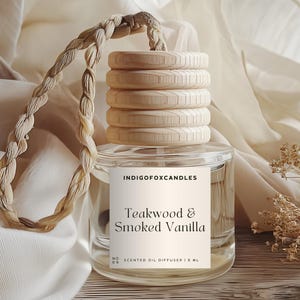 Op de afbeelding: Een heldere glazen geuroliediffuser met een houten dop en een gevlochten koord. Het label luidt "Teakwood & Smoked Vanilla" en "IndigoFoxCandles". De diffuser is 1,8 ml en staat op een houten oppervlak met een crèmekleurige stof op de achtergrond.