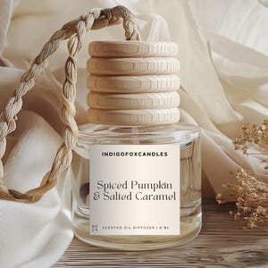 Könnte beinhalten: Ein klarer Glas-Duftöldiffusor mit Holzdeckel und geflochtenem Seilgriff. Auf dem Etikett steht "Spiced Pumpkin & Salted Caramel". Der Diffusor ist von einem cremefarbenen Stoff und Trockenblumen umgeben.