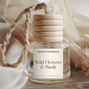 Könnte beinhalten: Ein klarer Glas-Duftöldiffusor mit einem Holzdeckel und einem geflochtenen Seil. Das Etikett lautet "Wild Flowers & Musk". Der Diffusor wird auf einer Holzoberfläche mit einem neutralfarbenen Stoffhintergrund präsentiert.