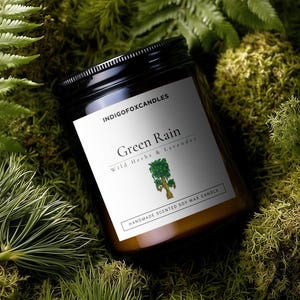 Op de afbeelding: Een glazen potkaars met een zwart deksel, met het label "Green Rain" met de tekst "Wild Herbs & Lavender". De kaars ligt op een bed van groen mos en varens. De tekst "HANDMADE SCENTED SOY WAX CANDLE" is ook aanwezig.