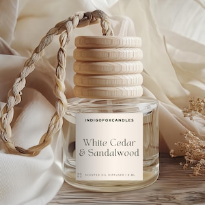 Könnte beinhalten: Ein klarer Glas-Duftöl-Diffusor mit einem Holzdeckel und einem geflochtenen Seil. Das Etikett lautet "White Cedar & Sandalwood". Der Diffusor wird auf einer Holzoberfläche mit weißem Stoffhintergrund und getrockneten Blumen präsentiert.