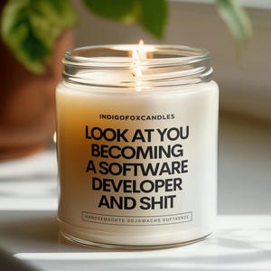 Op de afbeelding: Een brandende, witte sojawas kaars in een glazen pot. De kaars heeft zwarte tekst die luidt: "LOOK AT YOU BECOMING A SOFTWARE DEVELOPER AND SHIT". De kaars staat op een wit oppervlak met een groene plant op de achtergrond.