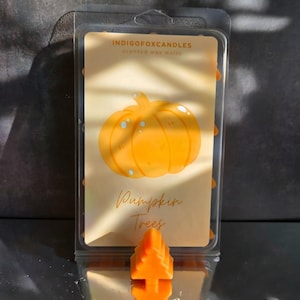 Könnte beinhalten: Eine transparente Kunststoffverpackung mit orangefarbenen Duftwachsschmelzen. Das Etikett zeigt eine Grafik eines orangefarbenen Kürbisses und die Worte "Pumpkin Trees". Der Markenname "INDIGOFoxCandles" ist ebenfalls sichtbar.