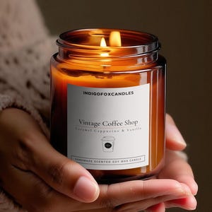 Könnte beinhalten: Eine brennende Kerze in einem braunen Glasgefäß mit einem weißen Etikett, das INDIGOFOXCANDLES, Vintage Coffee Shop, Caramel Cappuccino & Vanilla sowie ein kleines Kaffeetassen-Symbol zeigt. Das Etikett bezeichnet sie als handgefertigte Sojawachskerze.