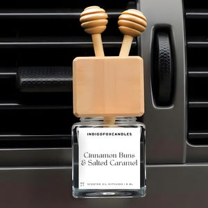 Könnte beinhalten: Ein Auto-Lufterfrischer mit einem Glasbehälter und Holzakzenten. Auf dem Etikett steht "Cinnamon Buns & Salted Caramel". Der Diffusor hat zwei hölzerne Honiglöffel-Aufsätze. Der Lufterfrischer befindet sich vor einer grauen Lüftungsöffnung.