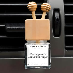 Può includere: Un profumatore per auto con un barattolo di vetro e dettagli in legno. L'etichetta recita "Red Apples & Cinnamon Sugar". Il diffusore ha due coperchi in legno con un design a cucchiaio per miele. Il profumatore è progettato per agganciarsi a una presa d'aria dell'auto.