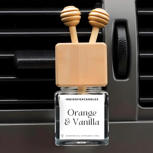 Könnte beinhalten: Ein Auto-Lufterfrischer mit einem Glasbehälter und Holzakzenten. Auf dem Etikett steht "Orange & Vanilla". Die Oberseite hat zwei hölzerne Honiglöffel-Diffusoren. Der Lufterfrischer ist für die Befestigung an einer Lüftungsöffnung im Auto konzipiert.