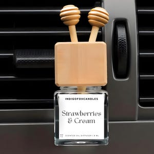 Può includere: Un profumatore per auto con un barattolo di vetro e dettagli in legno. L'etichetta recita "Strawberries & Cream". Il diffusore ha due coperchi in legno e un blocco di legno. Lo sfondo è una presa d'aria dell'auto.