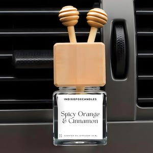 Könnte beinhalten: Ein Auto-Lufterfrischer mit einem Glasbehälter und Holzakzenten. Auf dem Etikett steht "Spicy Orange & Cinnamon". Der Diffusor hat zwei hölzerne Honiglöffel-Aufsätze. Der Lufterfrischer ist für die Befestigung an einer Lüftungsöffnung im Auto konzipiert.