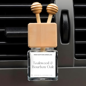 Könnte beinhalten: Ein Auto-Lufterfrischer mit einem Holzaufsatz und zwei dekorativen Holzkugeln. Das Glas hat ein Etikett mit der Aufschrift "Teakwood & Bourbon Oak" und "IndigoFoxCandles". Der Lufterfrischer befindet sich vor einer Lüftungsöffnung im Auto.