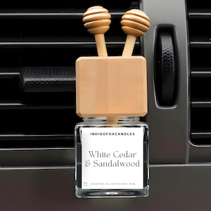 Può includere: Un deodorante per auto con un barattolo di vetro e dettagli in legno. L'etichetta recita "White Cedar & Sandalwood". La parte superiore presenta due diffusori in legno a forma di cucchiaio per il miele. Il deodorante è posto contro una presa d'aria grigia dell'auto.
