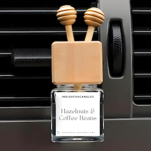 Könnte beinhalten: Ein Auto-Lufterfrischer mit einem Glasbehälter und Holzakzenten. Auf dem Etikett steht "Hazelnuts & Coffee Beans". Der Holzaufsatz hat zwei dekorative, honiglöffelartige Designs. Der Lufterfrischer ist für die Platzierung in einer Lüftungsöffnung im Auto konzipiert.