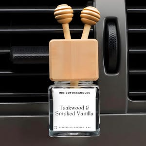 Op de afbeelding: Een auto-geurverspreider met een houten bovenkant en twee decoratieve houten ballen. De glazen fles heeft een label met de tekst "Teakwood & Smoked Vanilla" en "Indigo Fox Candles". De geurverspreider is ontworpen om op een ventilatierooster te klikken.