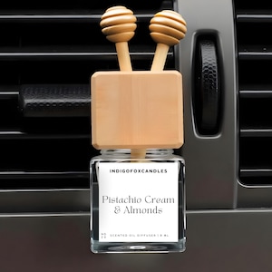 Könnte beinhalten: Ein Auto-Lufterfrischer mit einem Glasbehälter und Holzakzenten. Auf dem Etikett steht "Pistachio Cream & Almonds" und "Scented Oil Diffuser". Der Diffusor hat zwei kugelförmige Holzaufsätze.