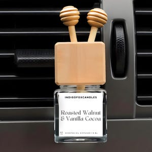 Könnte beinhalten: Ein Auto-Lufterfrischer mit einem Holzaufsatz und zwei dekorativen Holzkugeln. Das Glas hat ein Etikett mit der Aufschrift "Roasted Walnut & Vanilla Cocoa" und "IndigoFoxCandles". Der Lufterfrischer ist für die Platzierung in einer Lüftungsöffnung im Auto konzipiert.